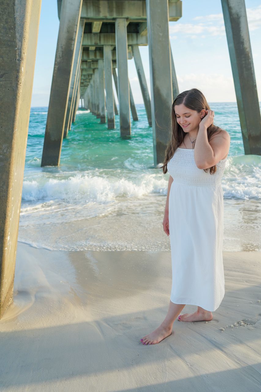 Brunette woman in white sundress posing barefoot under beach pier with ocean waves.