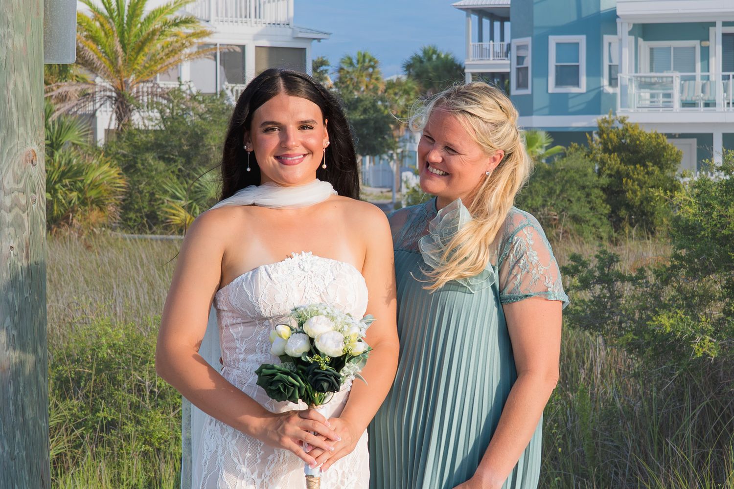 Bride in white lace dress holding white bouquet stands with bridesmaid in blue dress outdoors near beach.