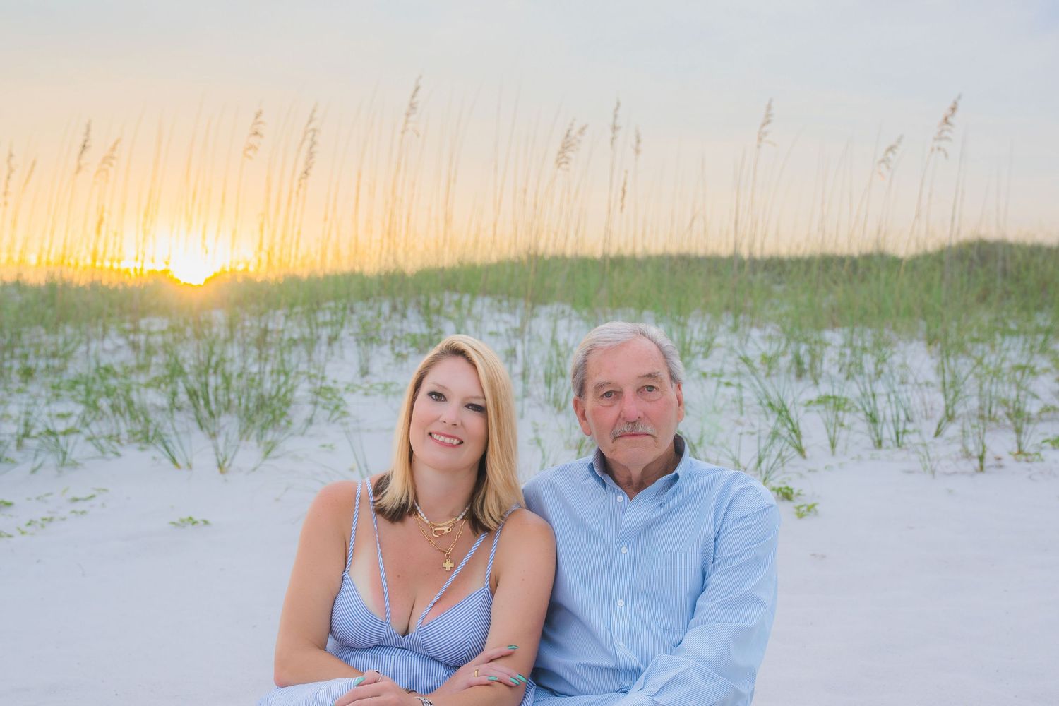 Couple sitting together on white sand beach at sunset with sea oats in background.