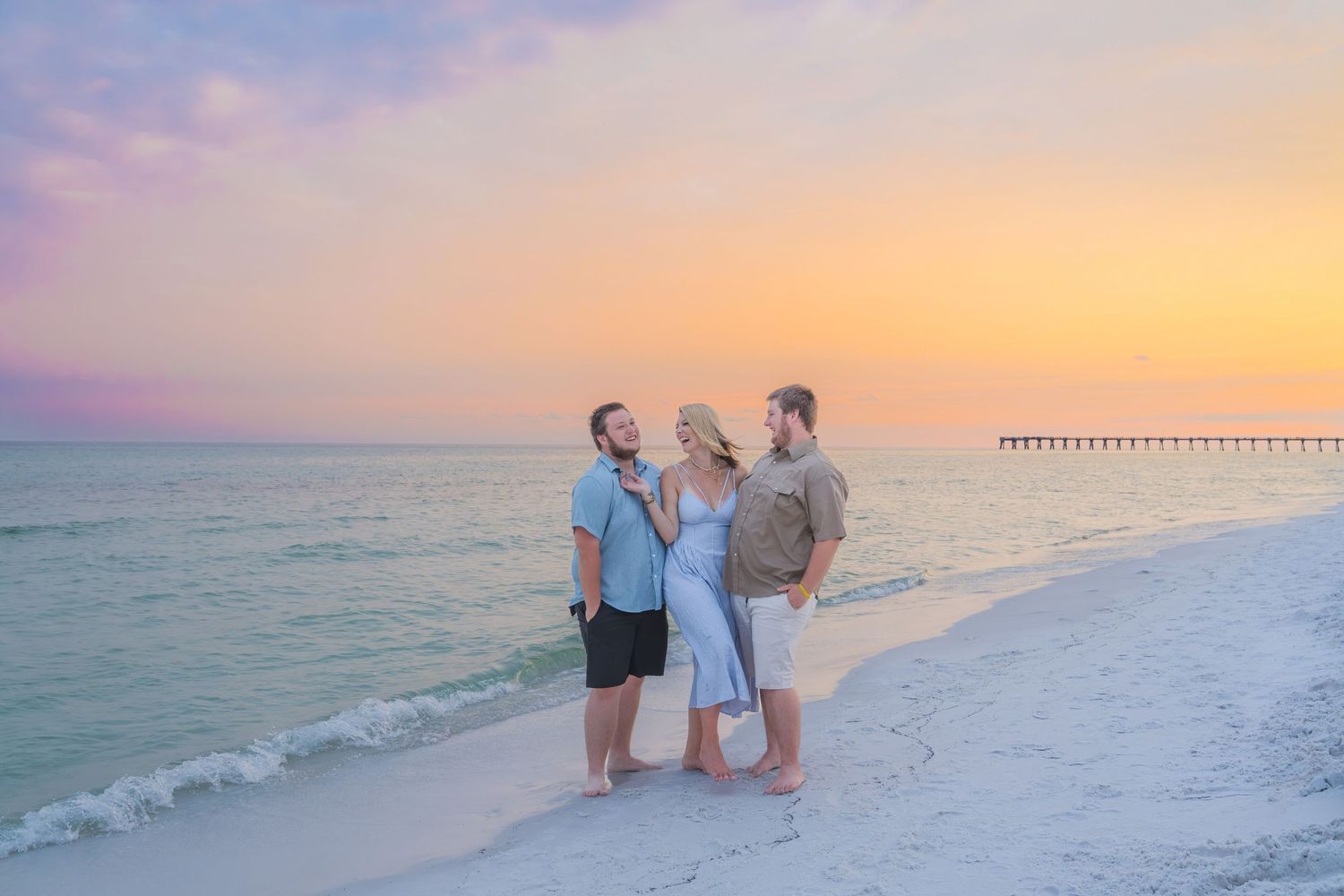 Family of three posing on a white sand beach at sunset with pastel pink and orange sky and ocean pier in background.
