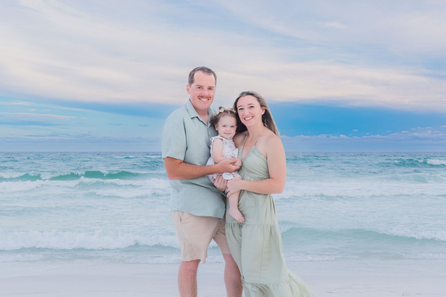 Family of three posing on a sandy beach with turquoise ocean waves and pastel sunset sky behind them.