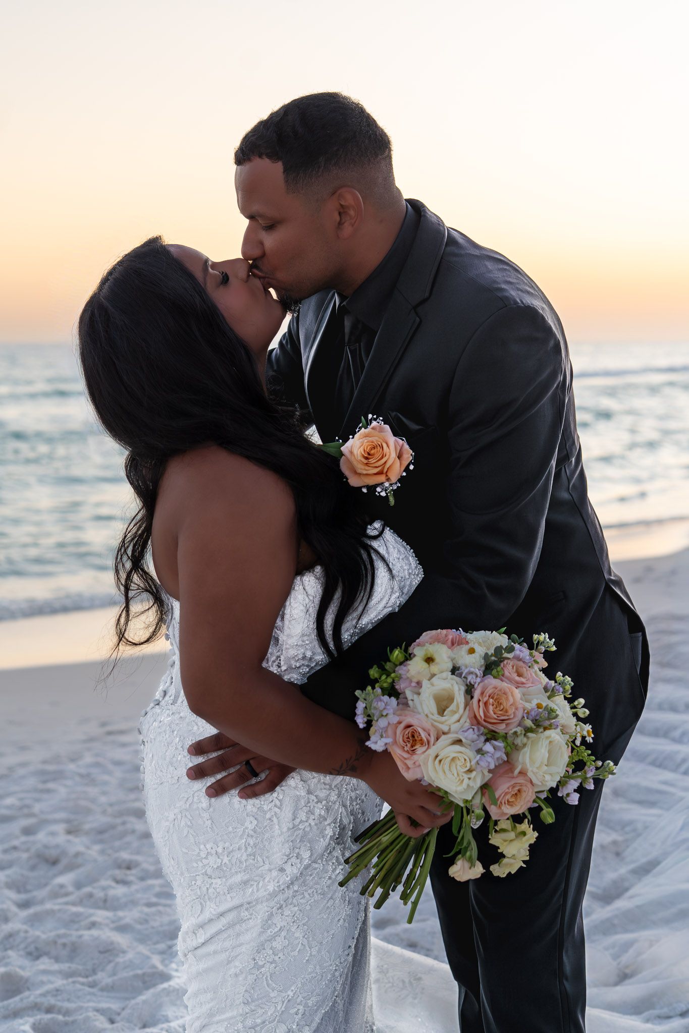 Couple sharing a romantic kiss on the beach at sunset, bride in white lace gown holding pastel rose bouquet, groom in black suit.