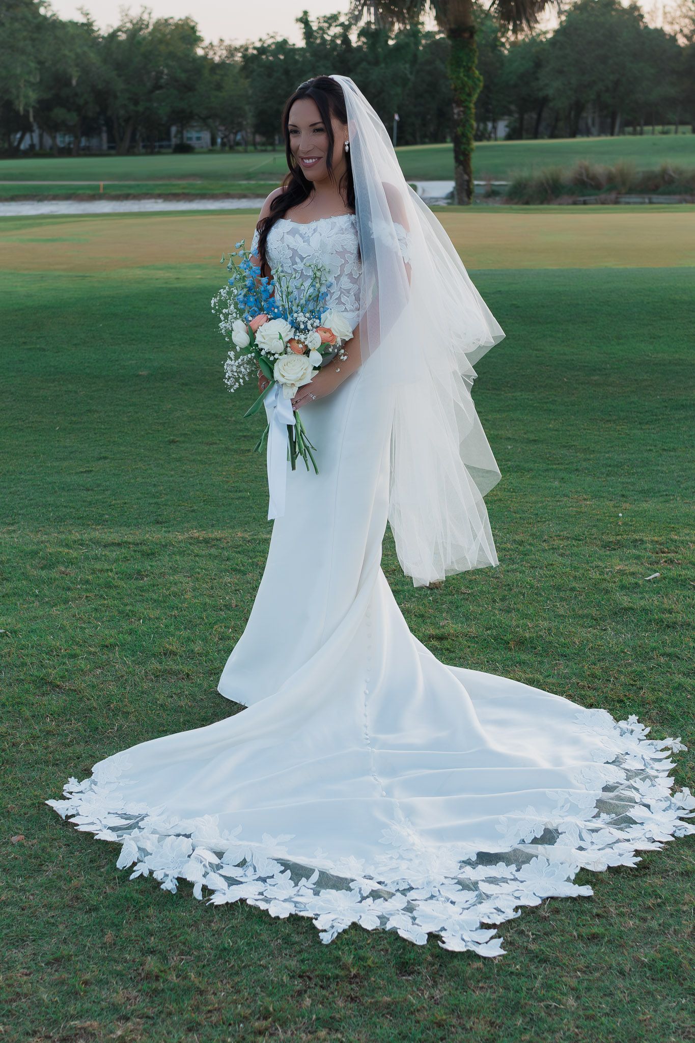 Bride in white mermaid gown with lace-trimmed veil and train holds blue and white bouquet on green lawn.