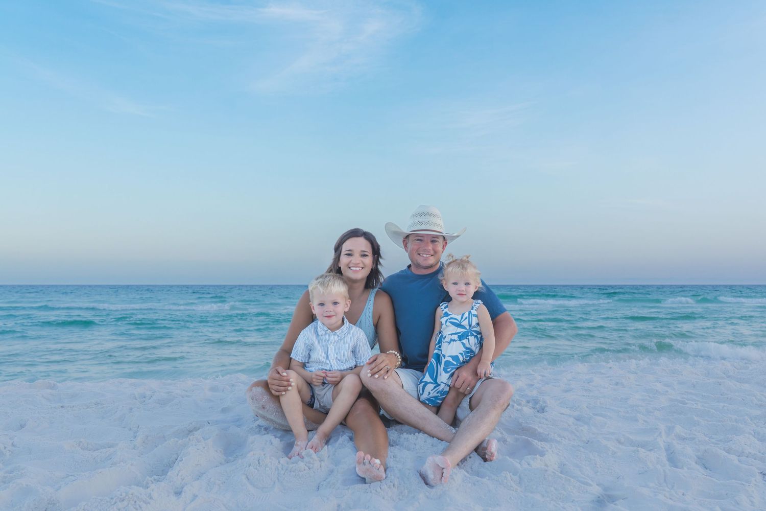 Family of four smiling together on a white sand beach with turquoise ocean water and blue sky backdrop.