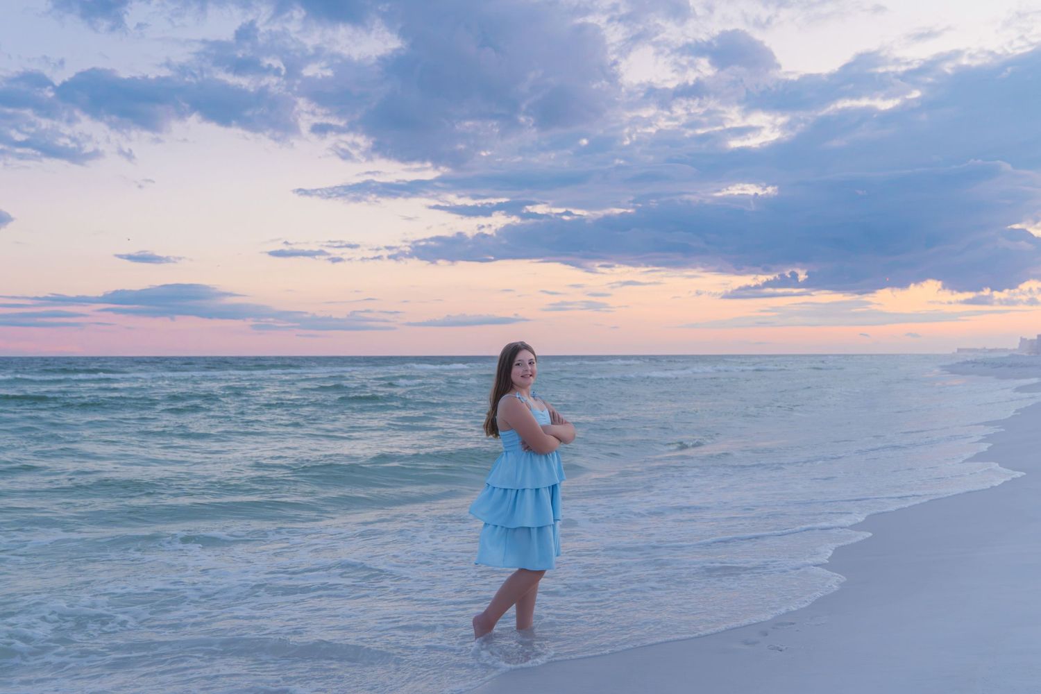 Young woman in a blue ruffled dress standing on a serene beach at sunset with colorful skies.