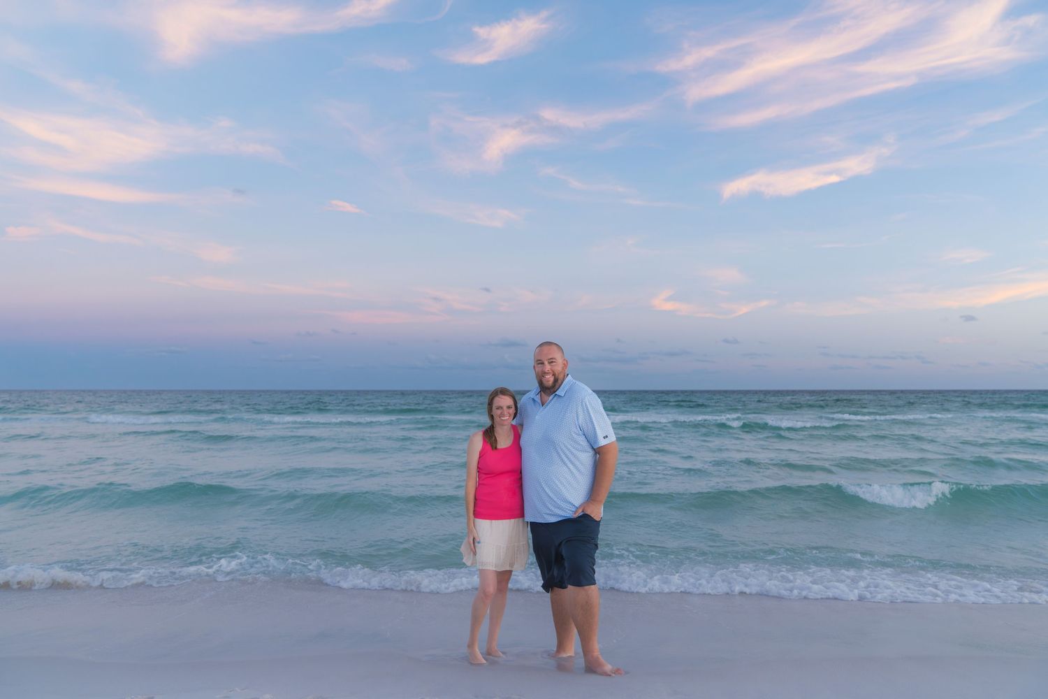 Couple standing together on a sandy beach at sunset with turquoise waves and pink sky.