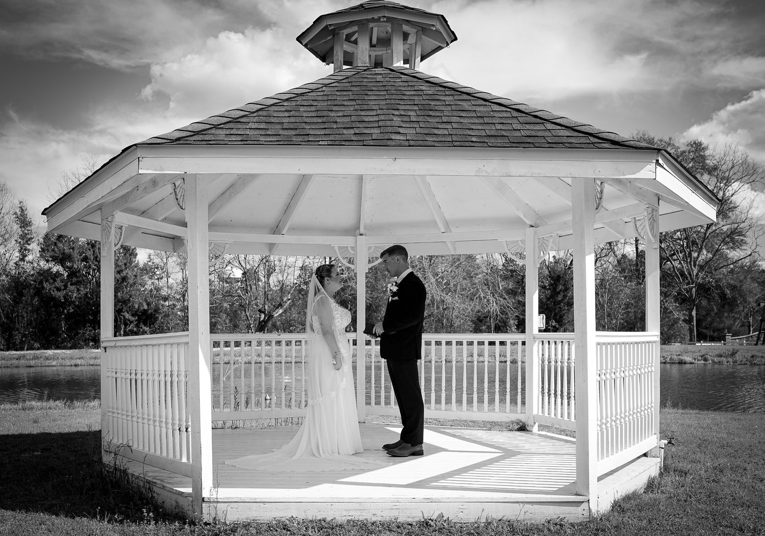Black and white photo of a bride and groom sharing a romantic moment inside a white gazebo at an outdoor wedding.