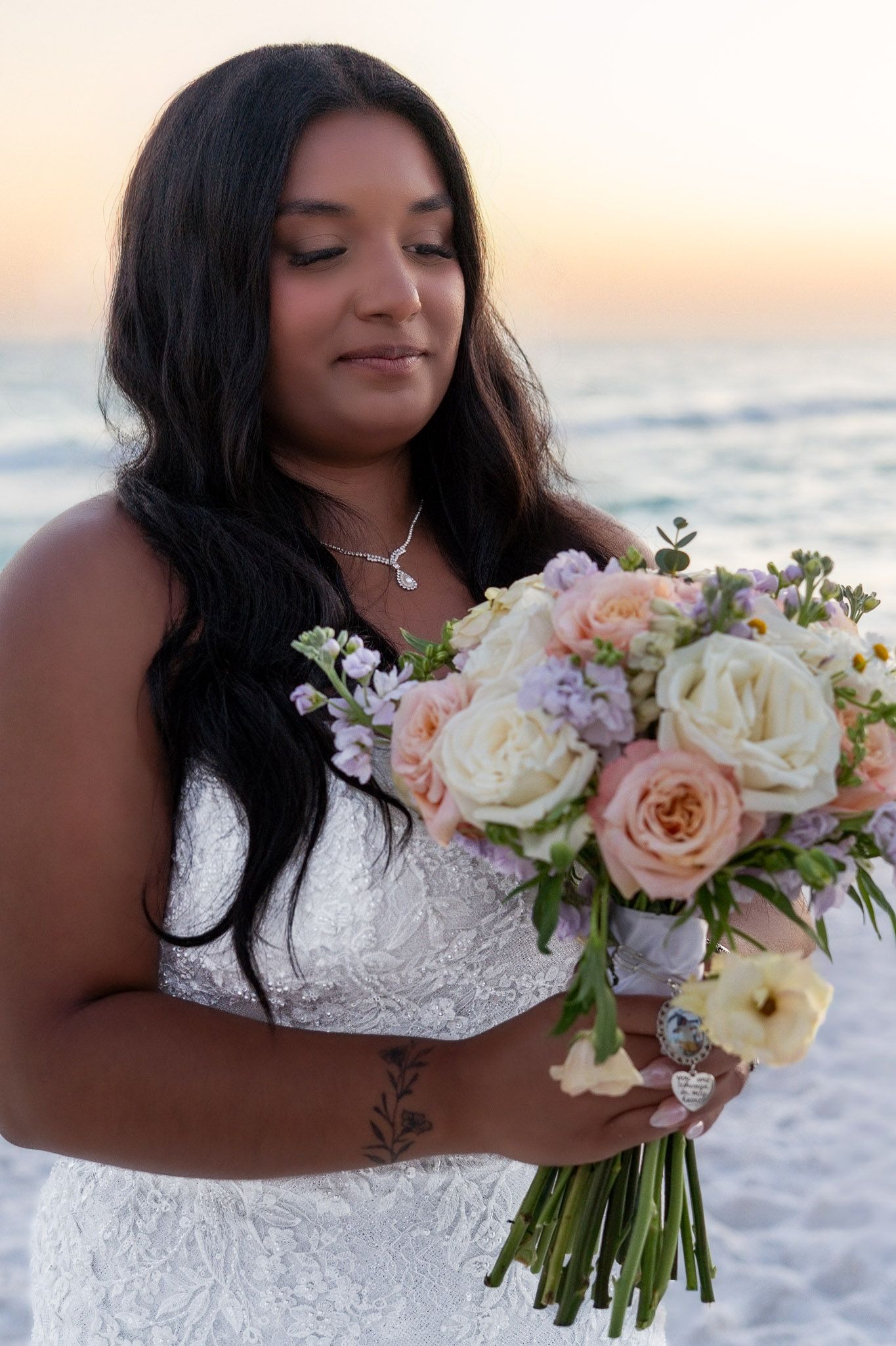 Bride in white lace gown holds pastel bouquet of roses on beach at sunset.