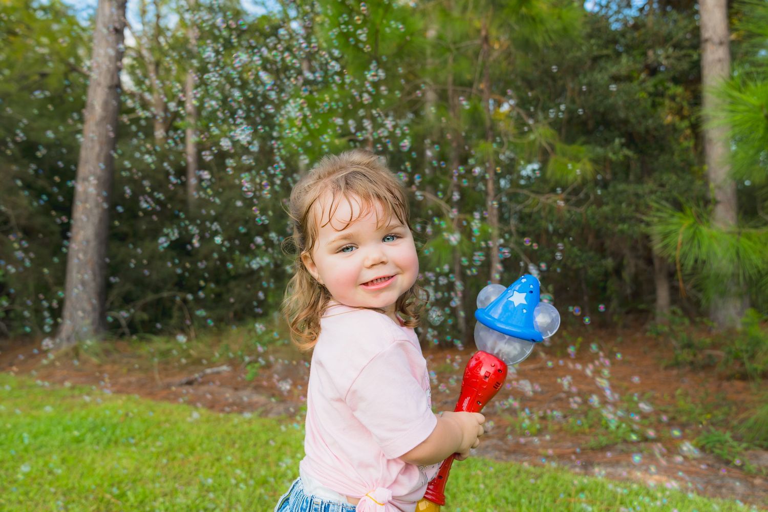 Toddler girl smiling outdoors holding a red and blue bubble wand toy with bubbles floating around her.