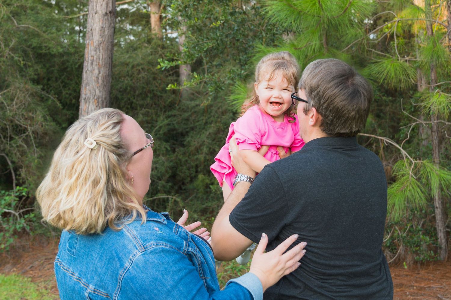 Two adults holding a laughing toddler girl in a pink dress outdoors among tall green pine trees.