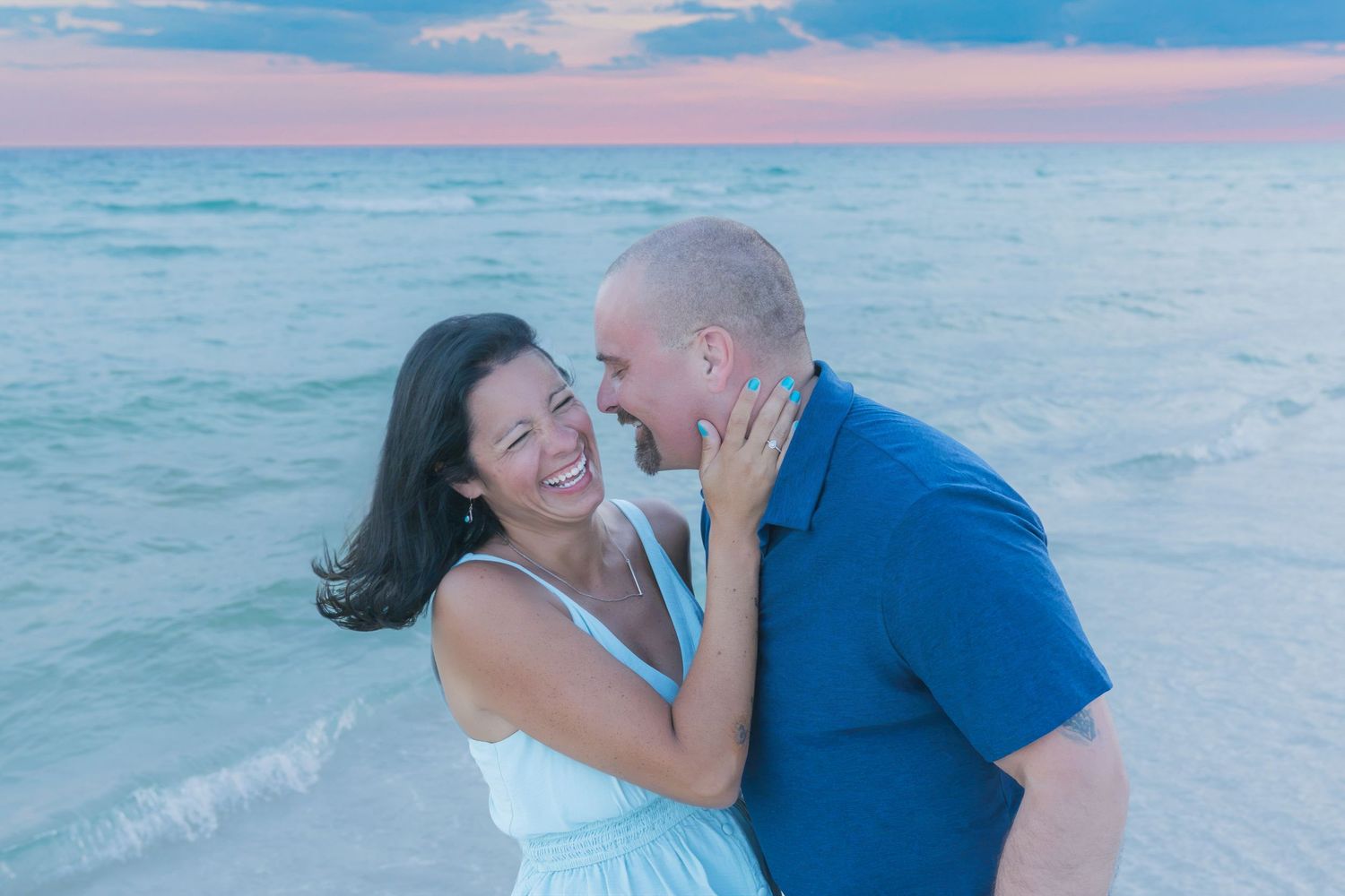 Couple laughing and embracing on a beach at sunset, woman in white dress touching man's face near ocean waves.