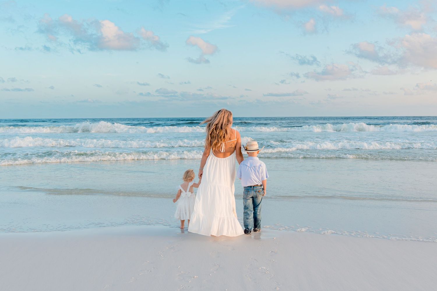 A mother in a white dress holds hands with two young children on a serene white sand beach at sunset.
