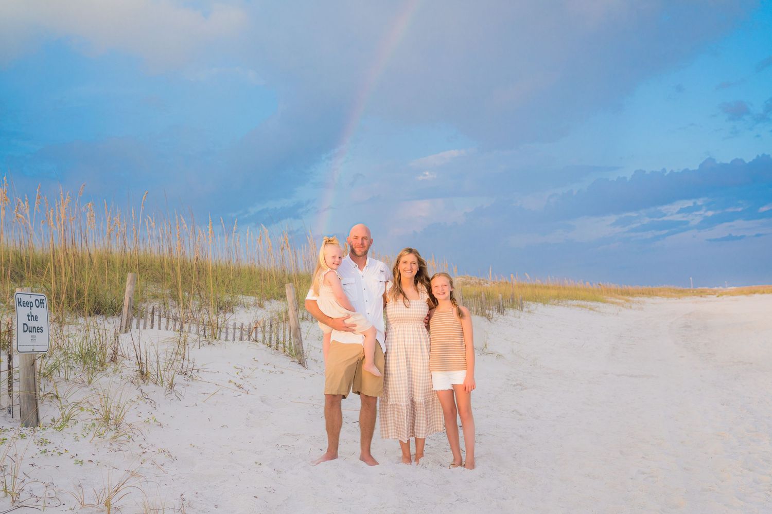 Family of four poses on white sand beach with rainbow and sea oats in background at golden hour.