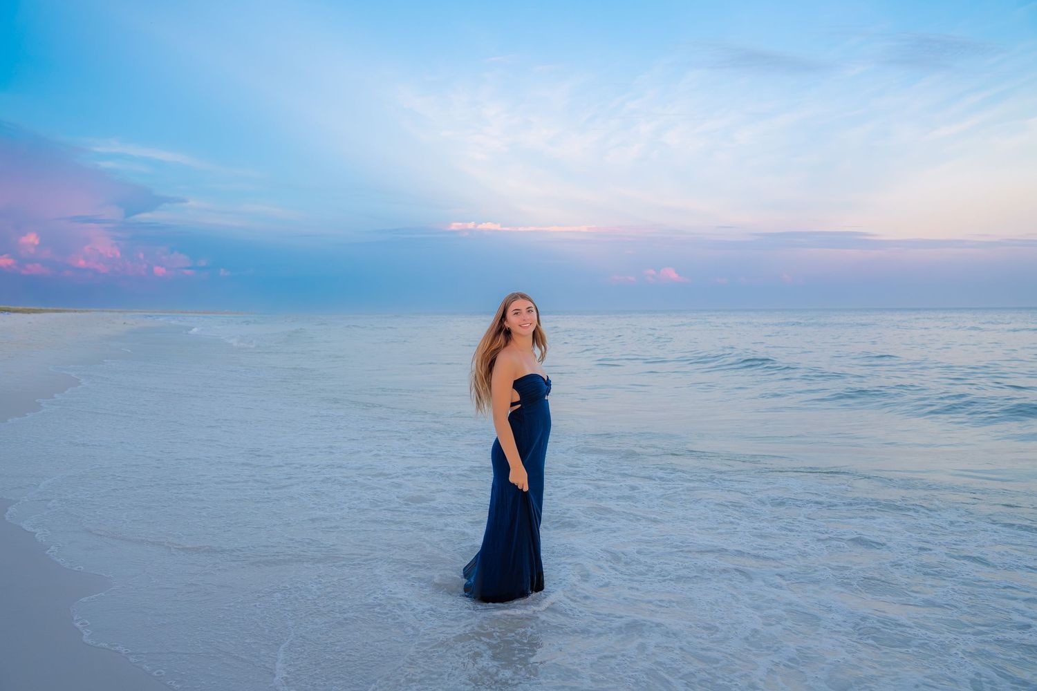 A woman in a navy blue gown stands on a serene beach at sunset with calm ocean waves.