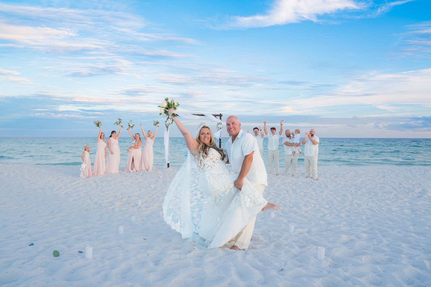 Bride and groom celebrate on white sand beach with wedding party in white attire behind them under blue sky.