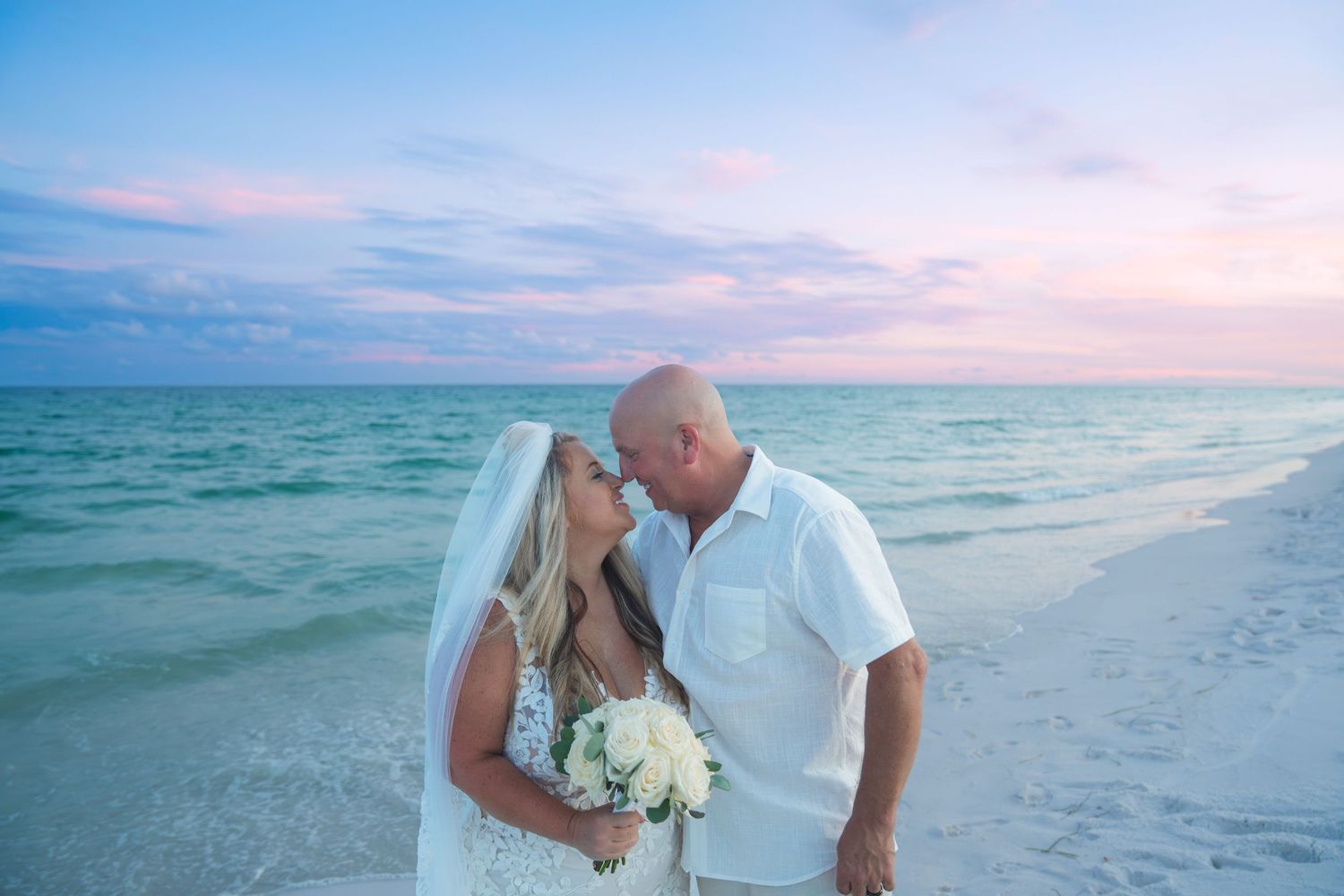 Bride and groom share a romantic kiss on a beach at sunset, with turquoise waters and a colorful sky behind them.