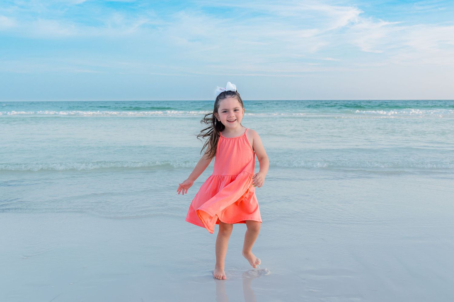Young girl in coral dress running joyfully on a sandy beach with turquoise ocean waves in the background.
