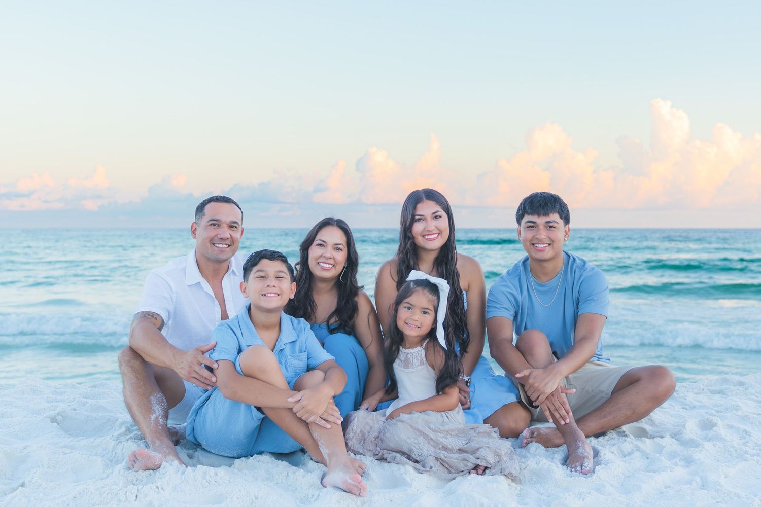 Family of six smiling together on a white sand beach at sunset with turquoise ocean waves in the background.