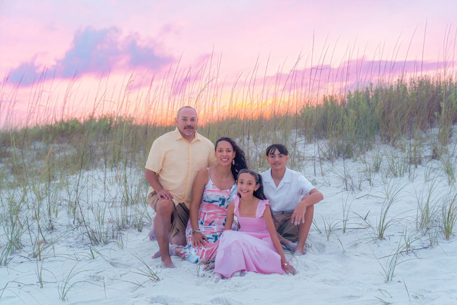 Family of four posing together on white sand dunes at sunset with pink and purple sky backdrop.