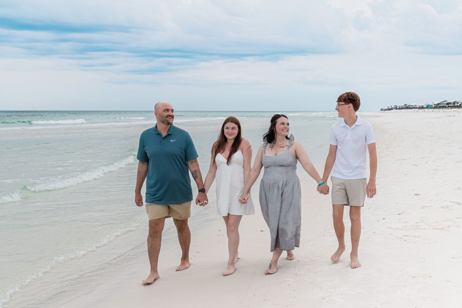 Family of four walking hand-in-hand on a white sand beach with turquoise ocean waves in the background.
