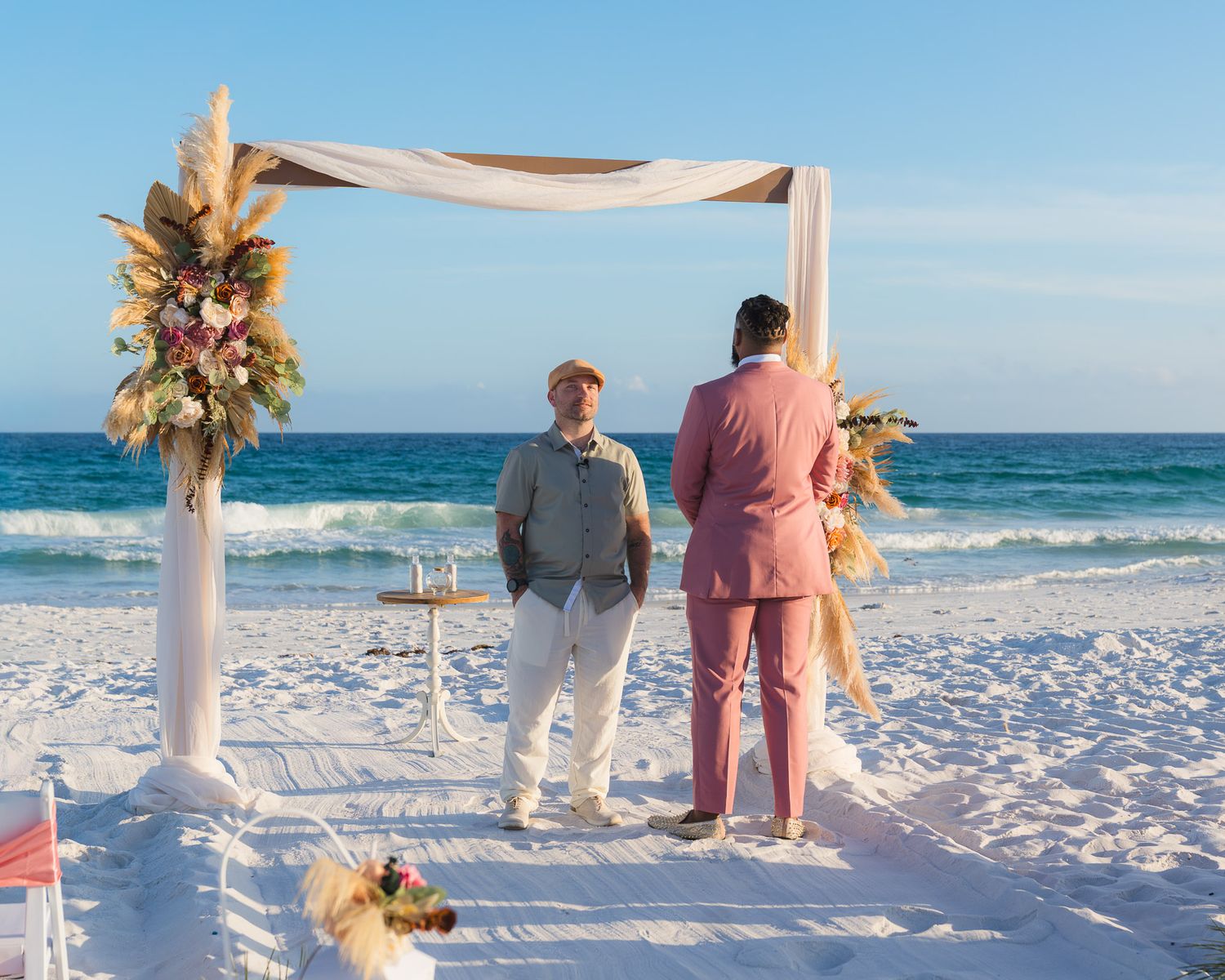Couple exchanging vows at a beachside altar with white draping and floral decor.
