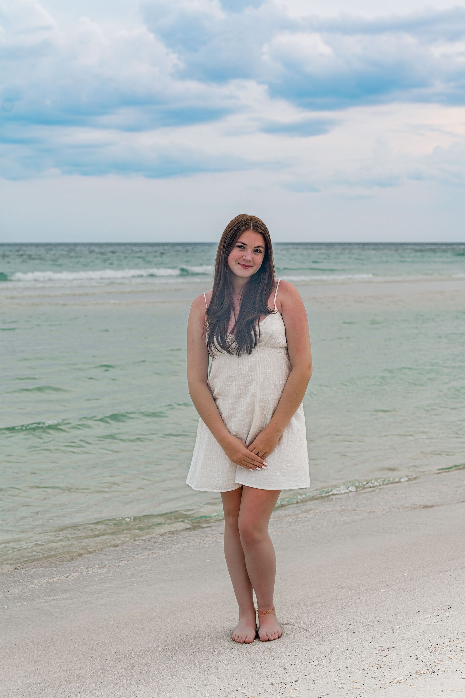 Young woman in white dress standing barefoot on a white sandy beach with turquoise ocean waves behind her.