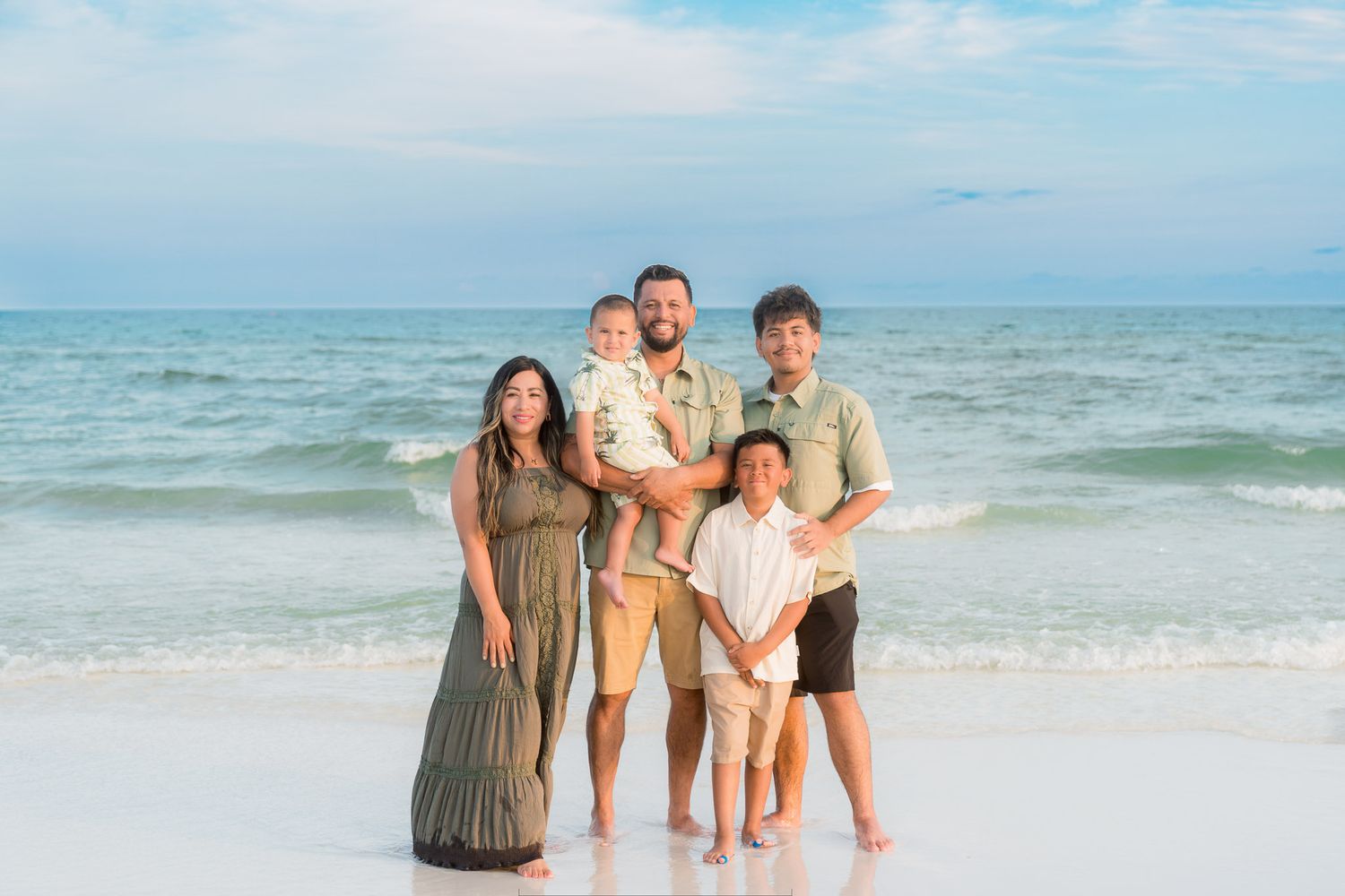 Family of five smiling together on a sandy beach with turquoise ocean waves in the background.