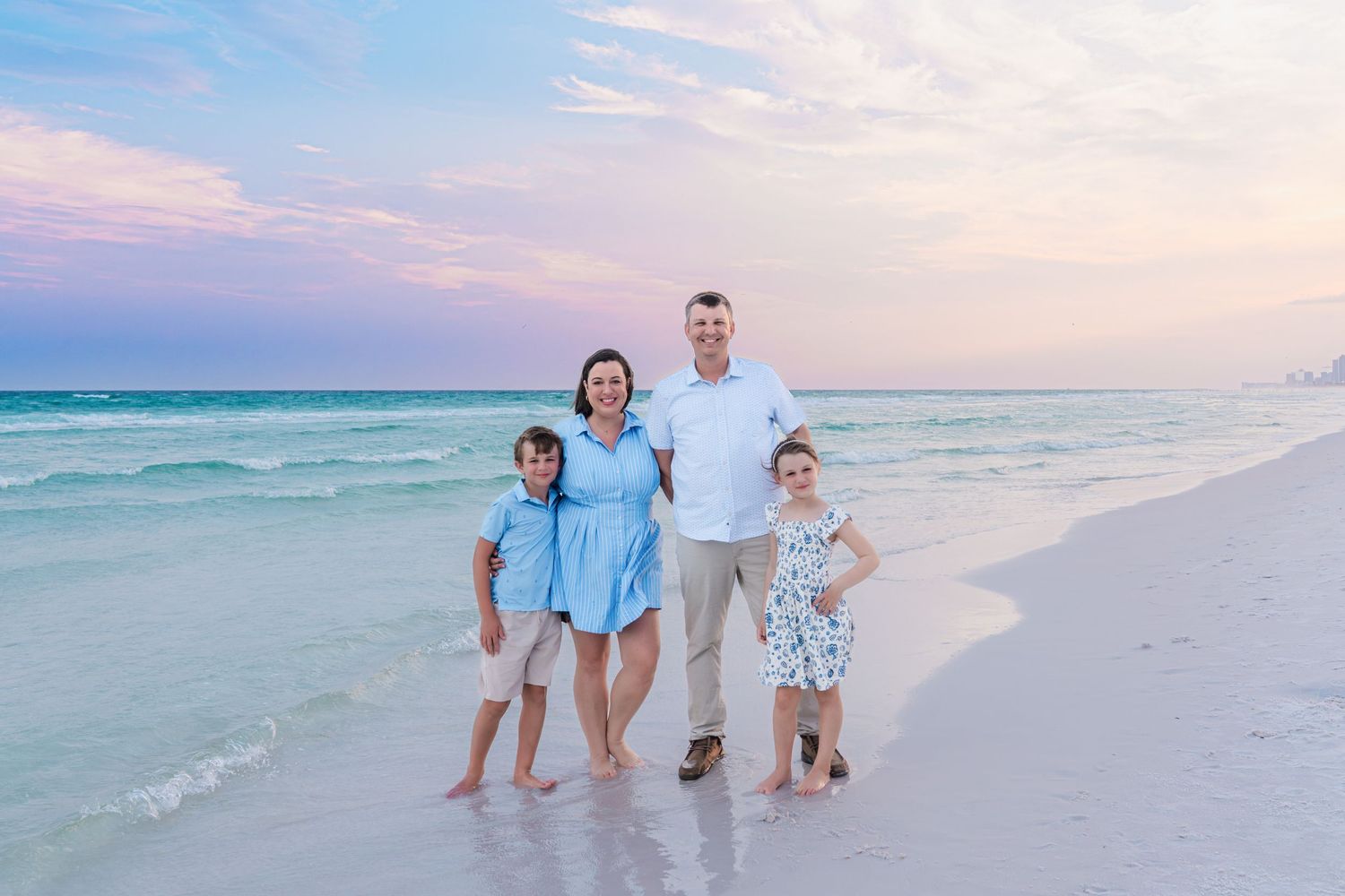 Family of four posing on a white sand beach at sunset with turquoise water and pink sky backdrop.