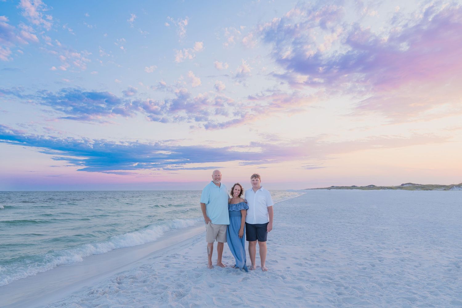Family of three posing on white sand beach at sunset with pastel pink and blue sky over calm ocean waters.