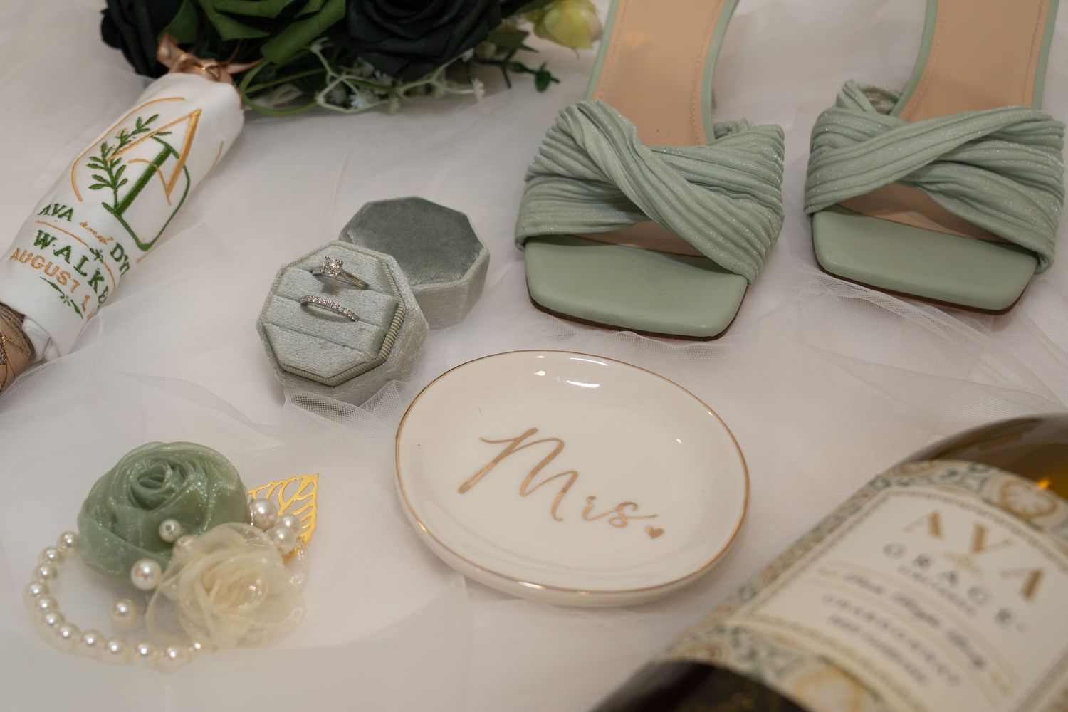 Sage green wedding flat lay with bridal heels, ring box, Mrs. dish, pearl corsage, and bouquet on white fabric.