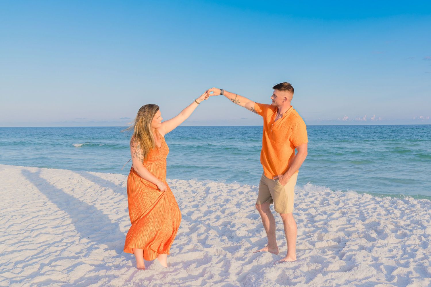Couple in matching orange outfits dancing on a white sand beach with turquoise ocean water in the background.