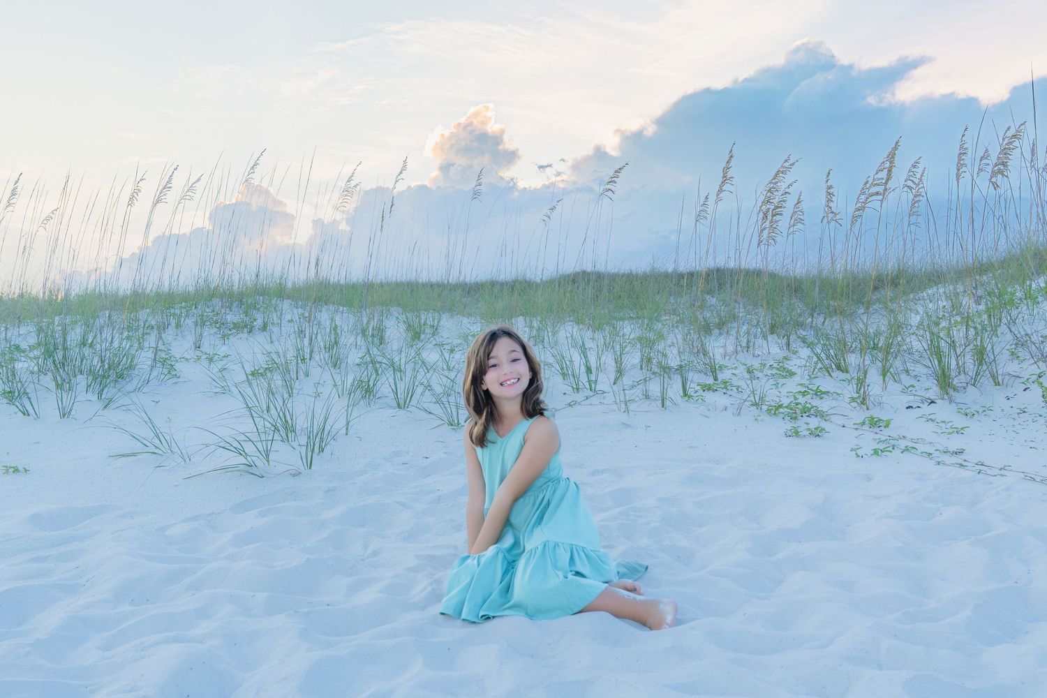 A young girl in a teal dress sits on white sand dunes with sea oats and clouds at a serene beach.