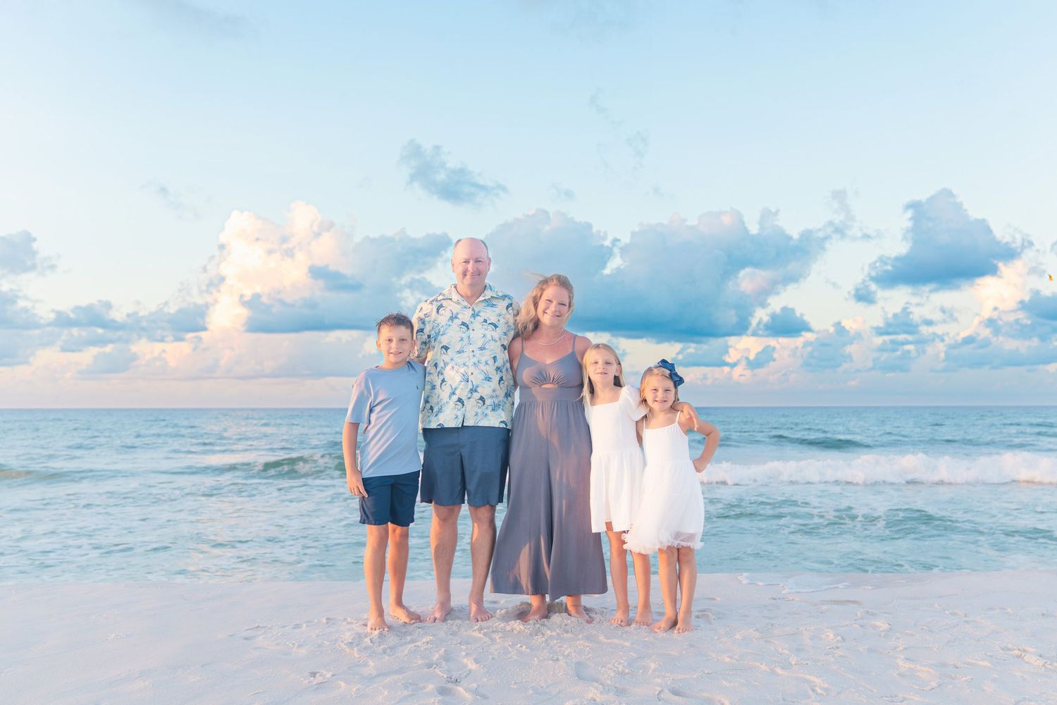 Family of five smiling together on a sandy beach at sunset with ocean waves and pastel clouds behind them.