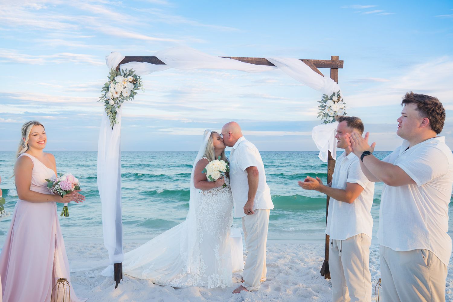 Couple shares first kiss at beach wedding ceremony under floral arch with ocean backdrop.