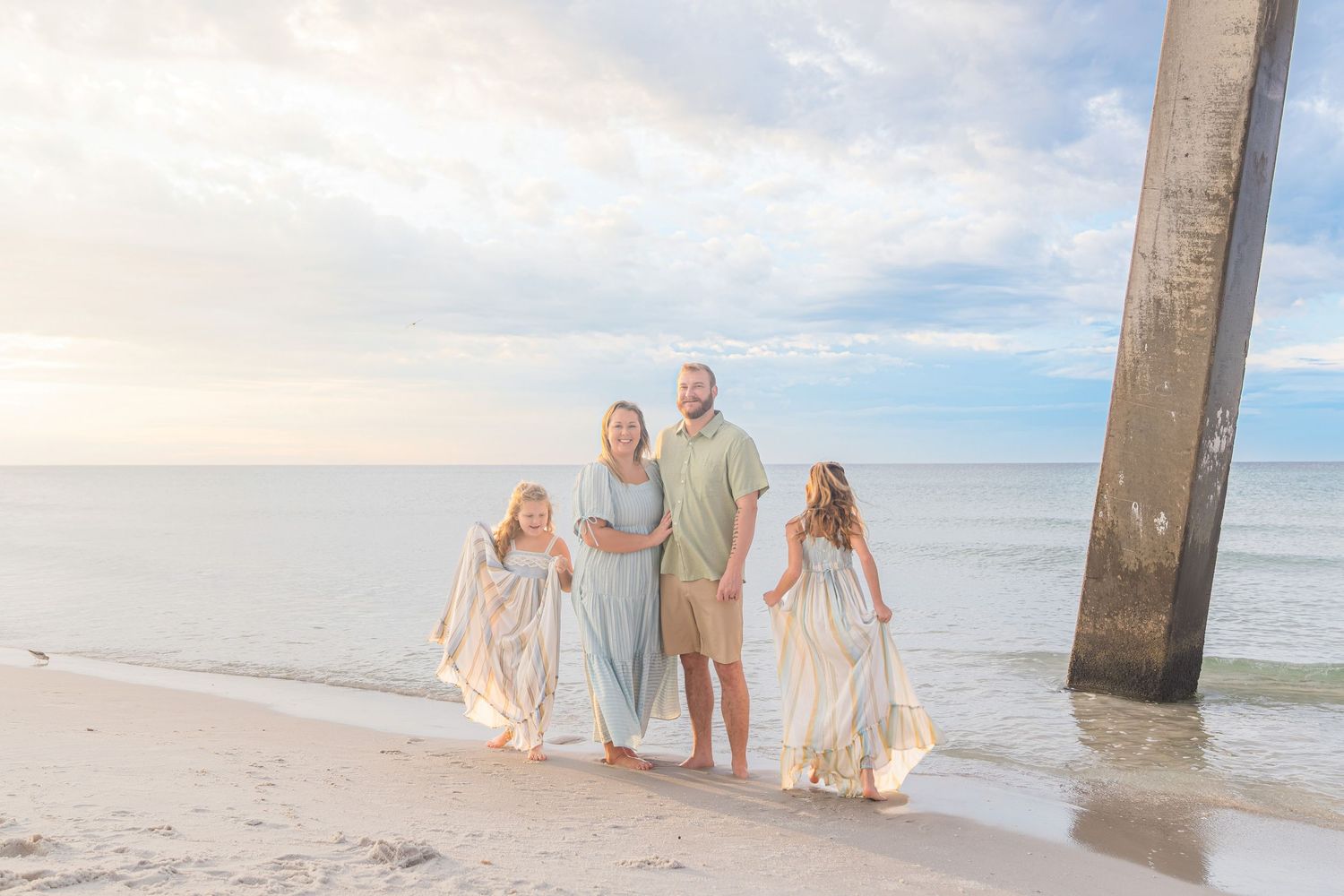 Family of four posing on a sandy beach near a pier at sunset, wearing flowing pastel outfits with ocean waves behind them.