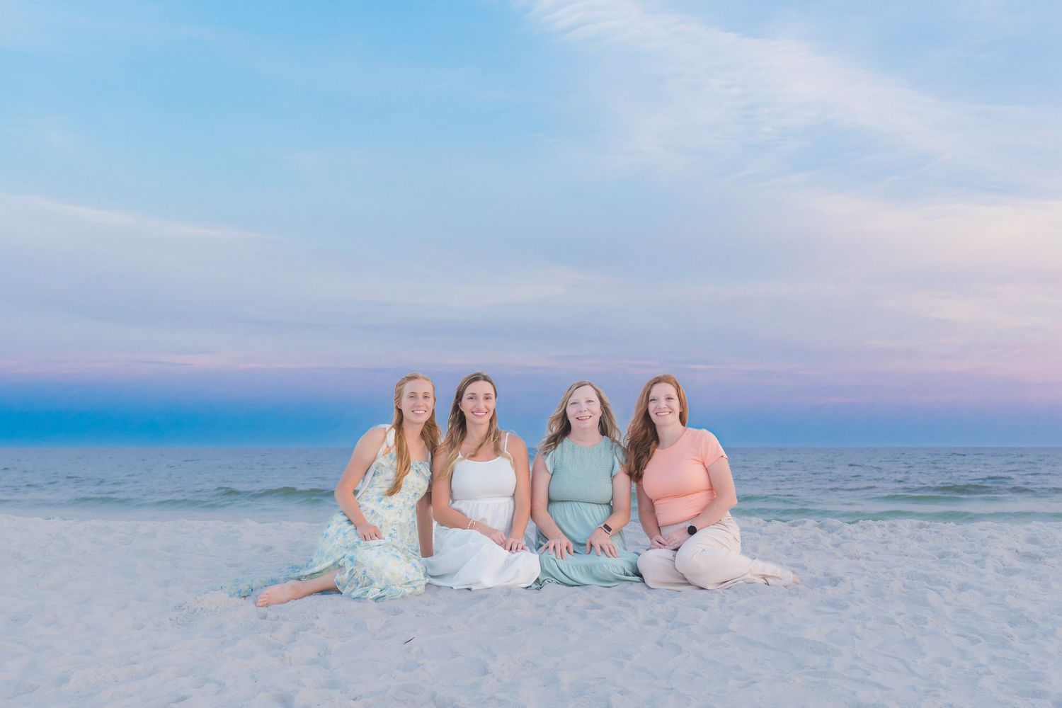 Four women sitting together on a white sand beach at sunset with pastel pink and blue sky backdrop.