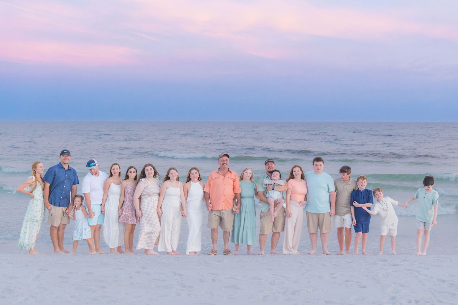 Large family group portrait on a sandy beach at sunset with pastel pink and blue sky backdrop.