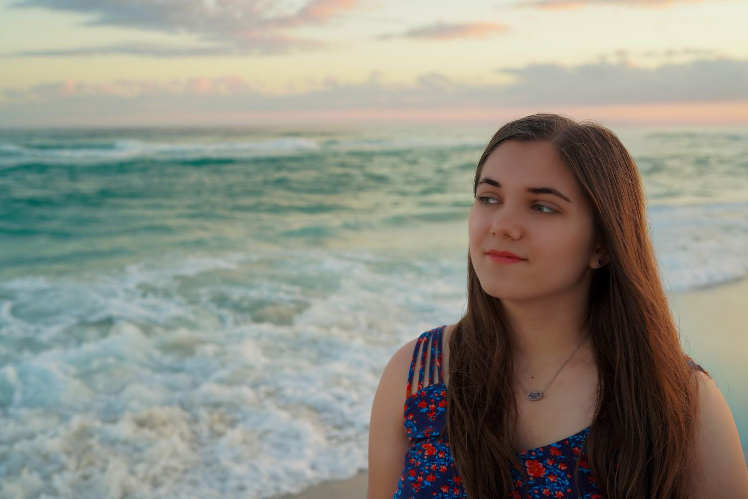 Young woman with long brown hair gazing at ocean waves during colorful beach sunset.