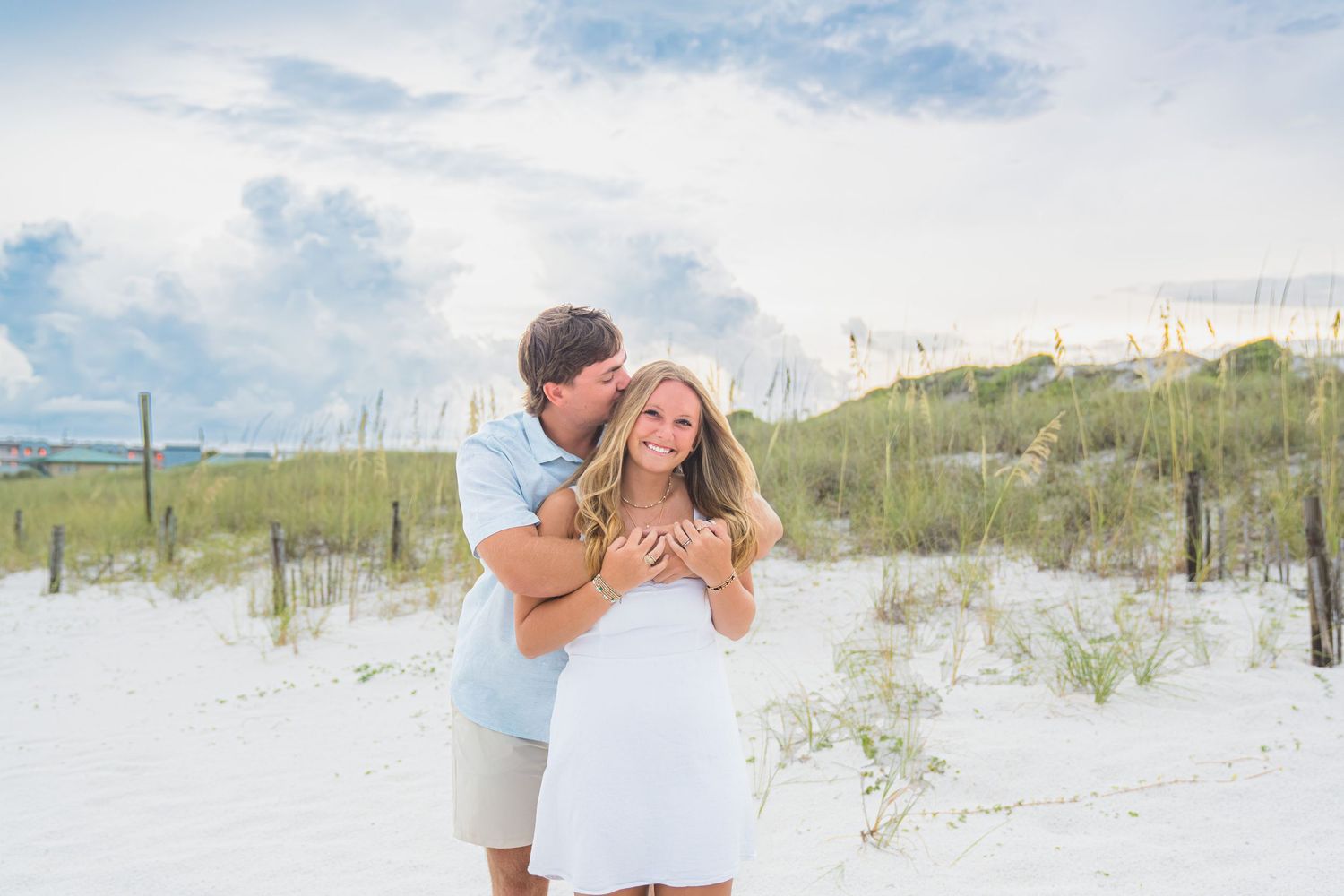 Couple embracing on white sand beach dunes, woman in white dress, man in blue shirt, cloudy sky backdrop.