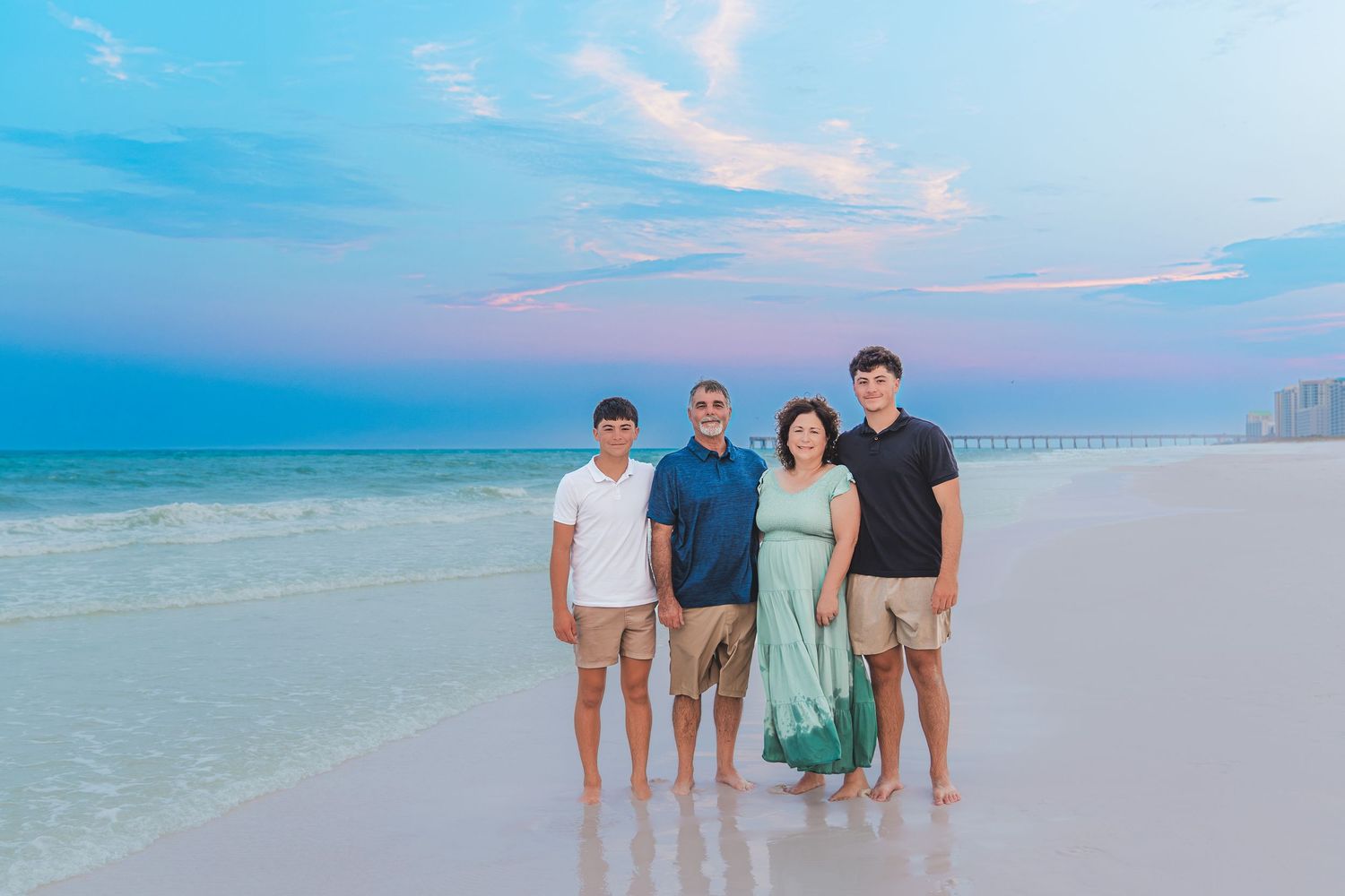 Family of four posing on a white sand beach at sunset with turquoise water and pastel sky.