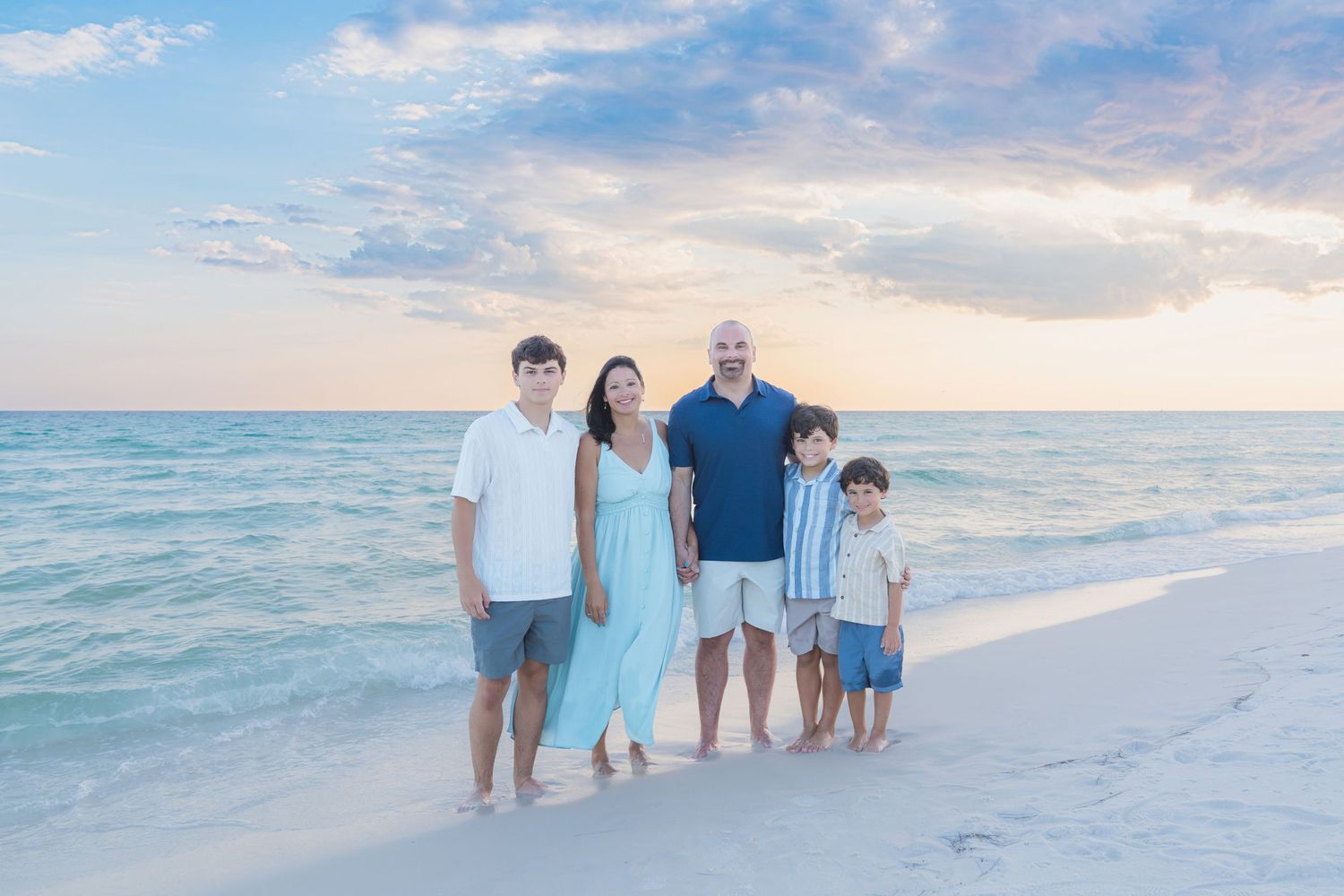 Family of five posing on a white sand beach at sunset with turquoise ocean waves in the background.