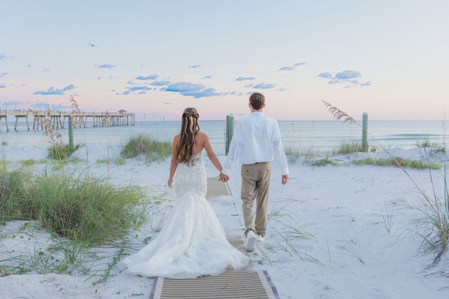 Bride and groom walking hand-in-hand on a beach boardwalk at sunset with a pier in the background.
