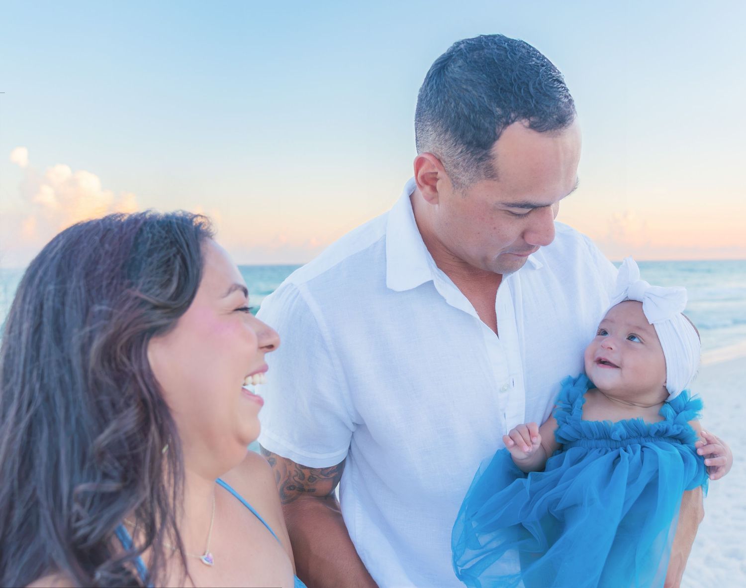 Happy family with parents smiling at their baby girl in a blue dress on a sunny beach at sunset.