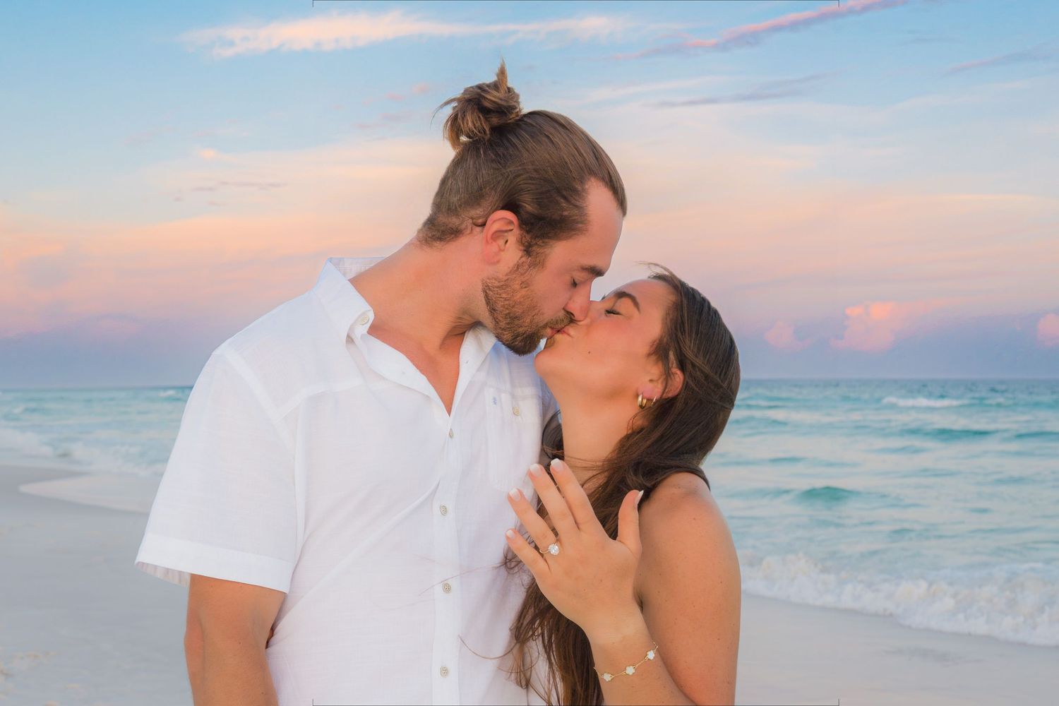 Couple kissing on a beach at sunset, woman showing engagement ring, wearing white outfits with ocean backdrop.