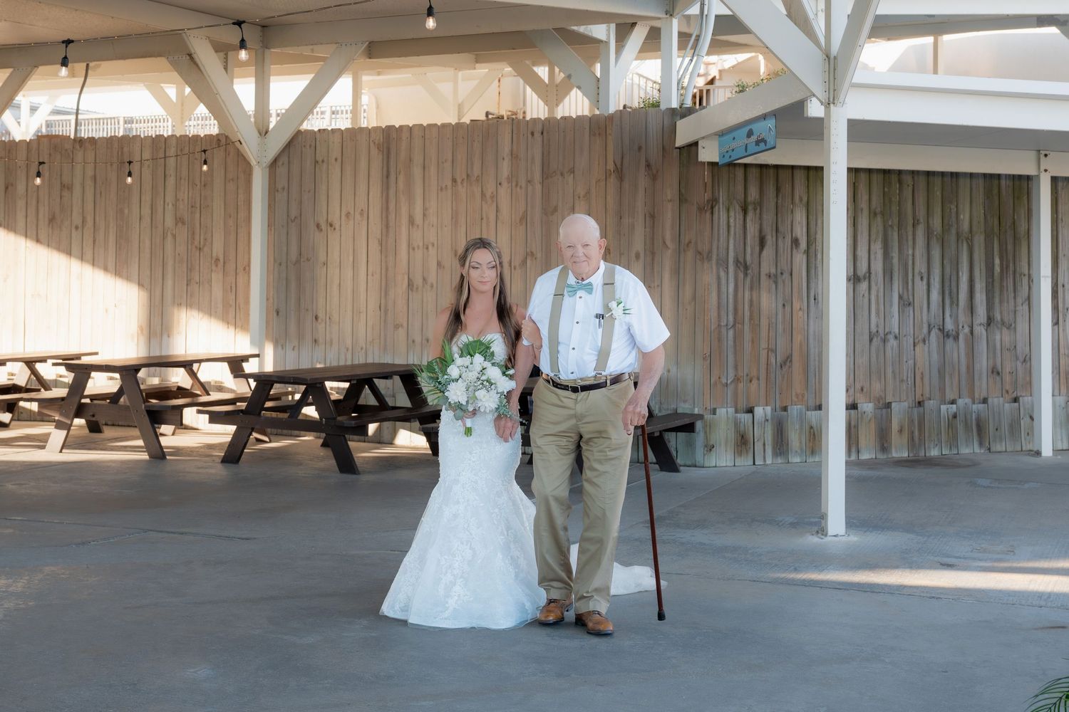 A bride in a white mermaid gown poses with an elderly man holding a cane at an outdoor venue.