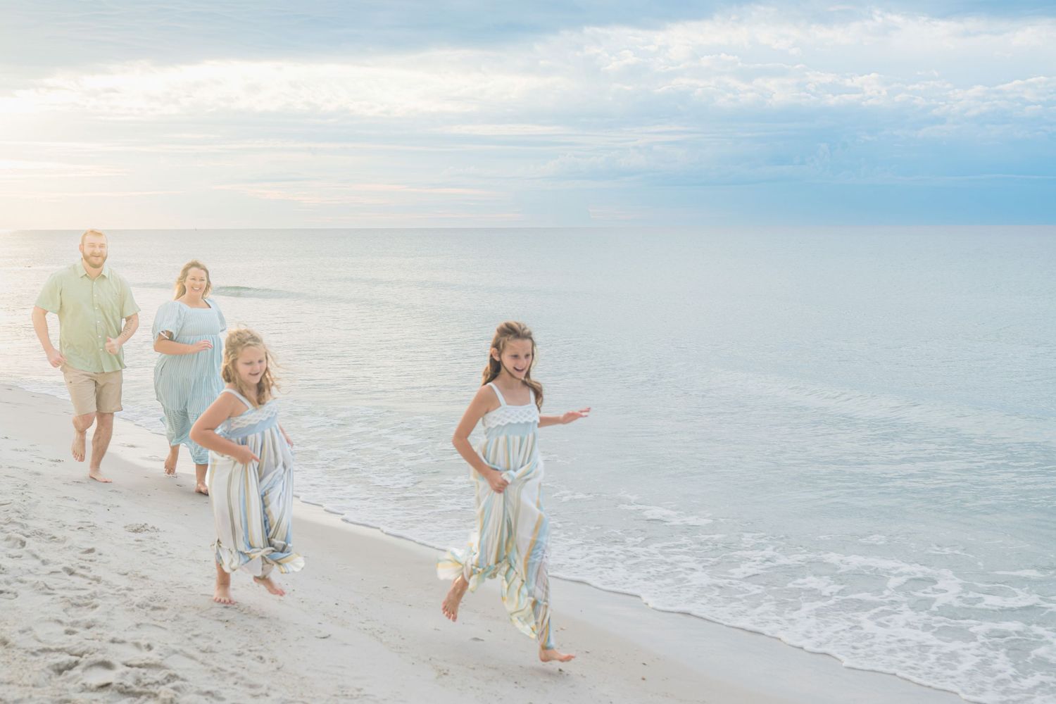 Family of four running and laughing on a sandy beach at sunset, wearing light blue and green outfits.