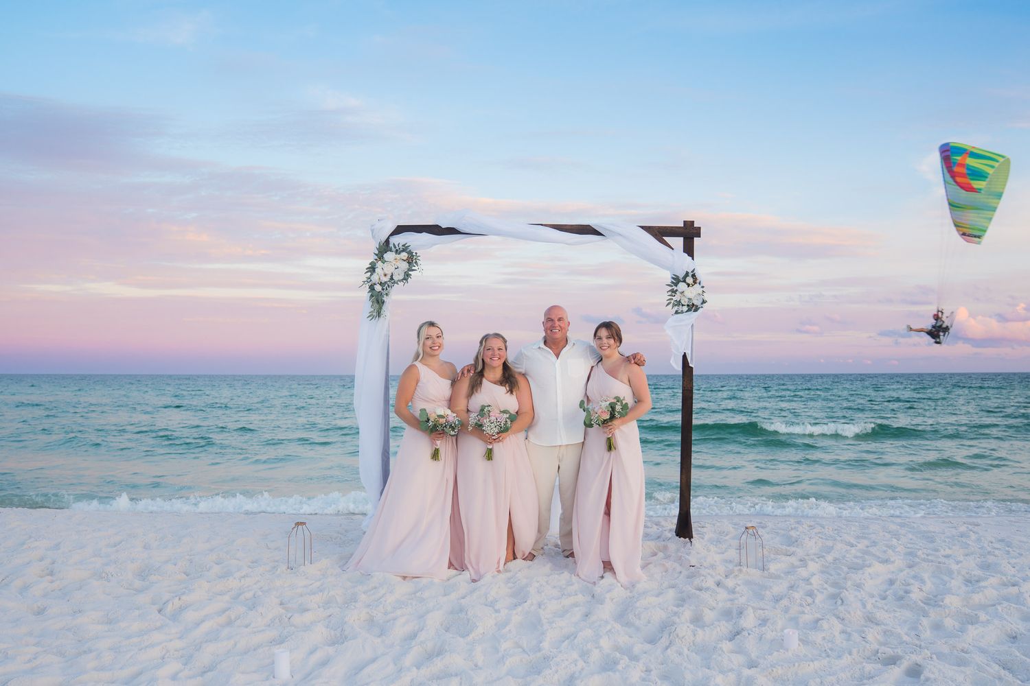 Beach wedding ceremony at sunset with bride, groom, and two bridesmaids in pink dresses under a white floral arch.