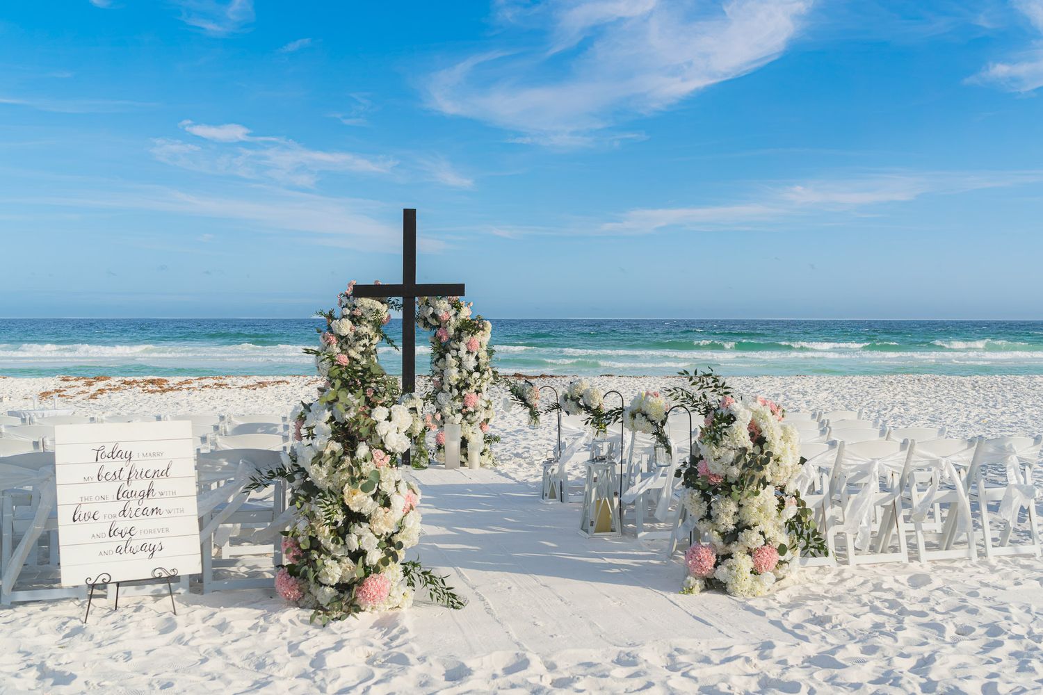 Elegant beach wedding setup with floral arch, wooden cross, white chairs on white sand with ocean backdrop.