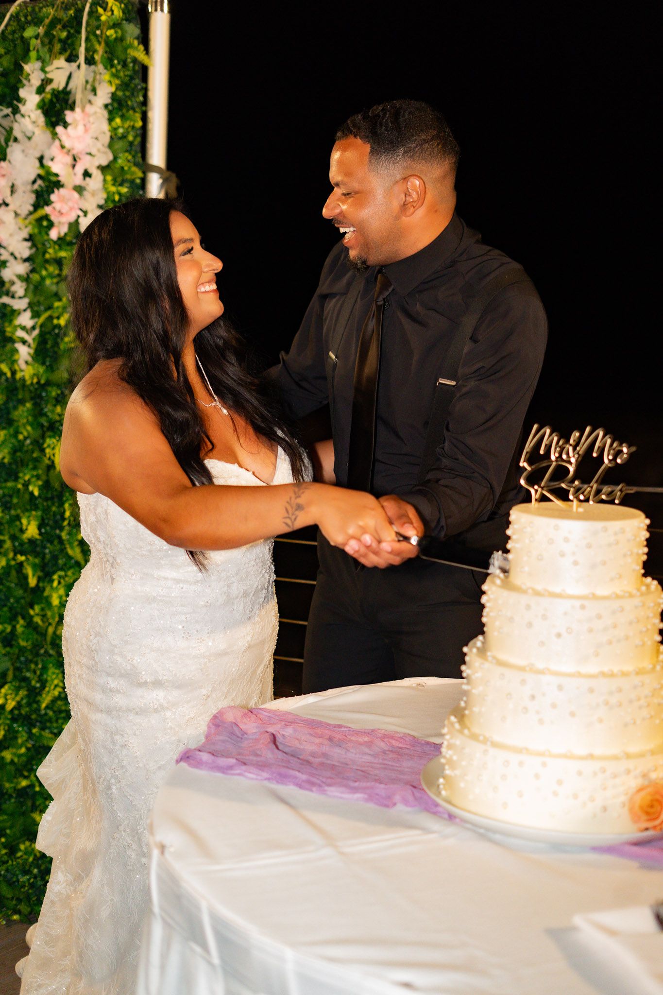 Bride and groom laugh together while cutting their four-tier white wedding cake at the evening reception.