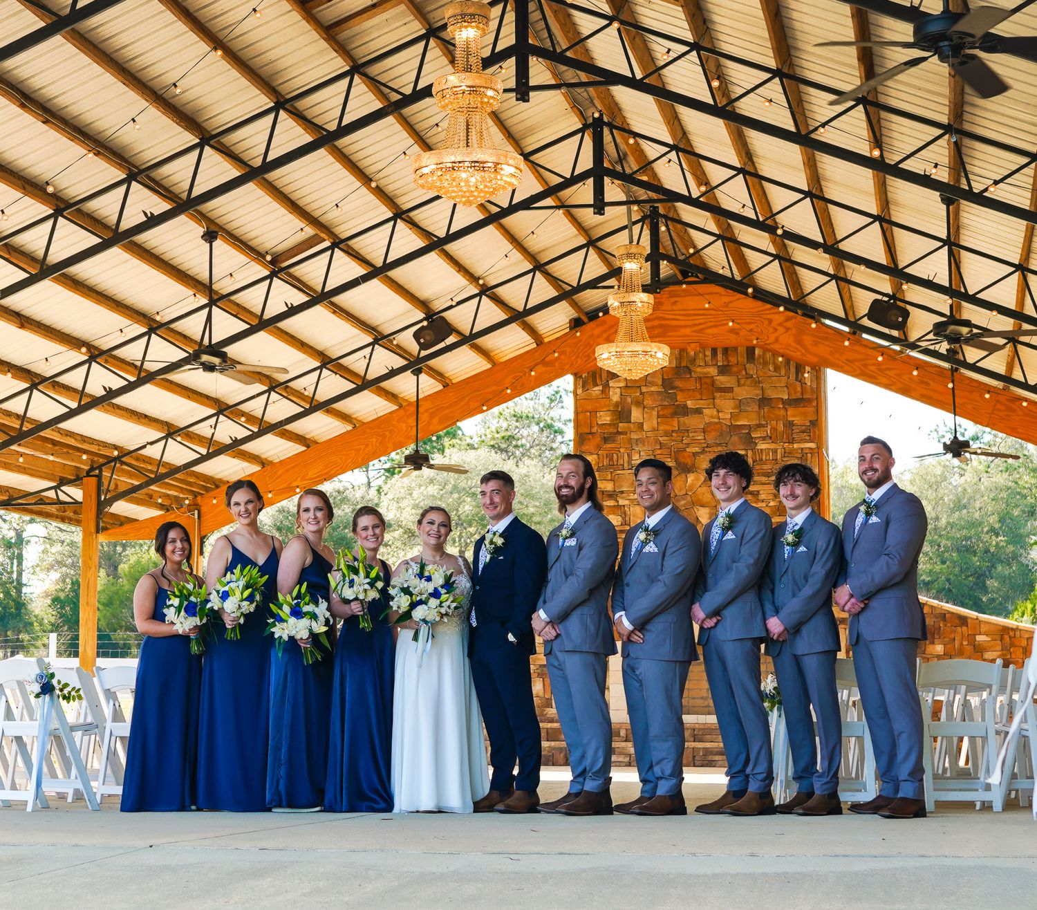 Full wedding party in navy and grey attire pose together inside rustic chandelier-lit wedding venue pavilion.
